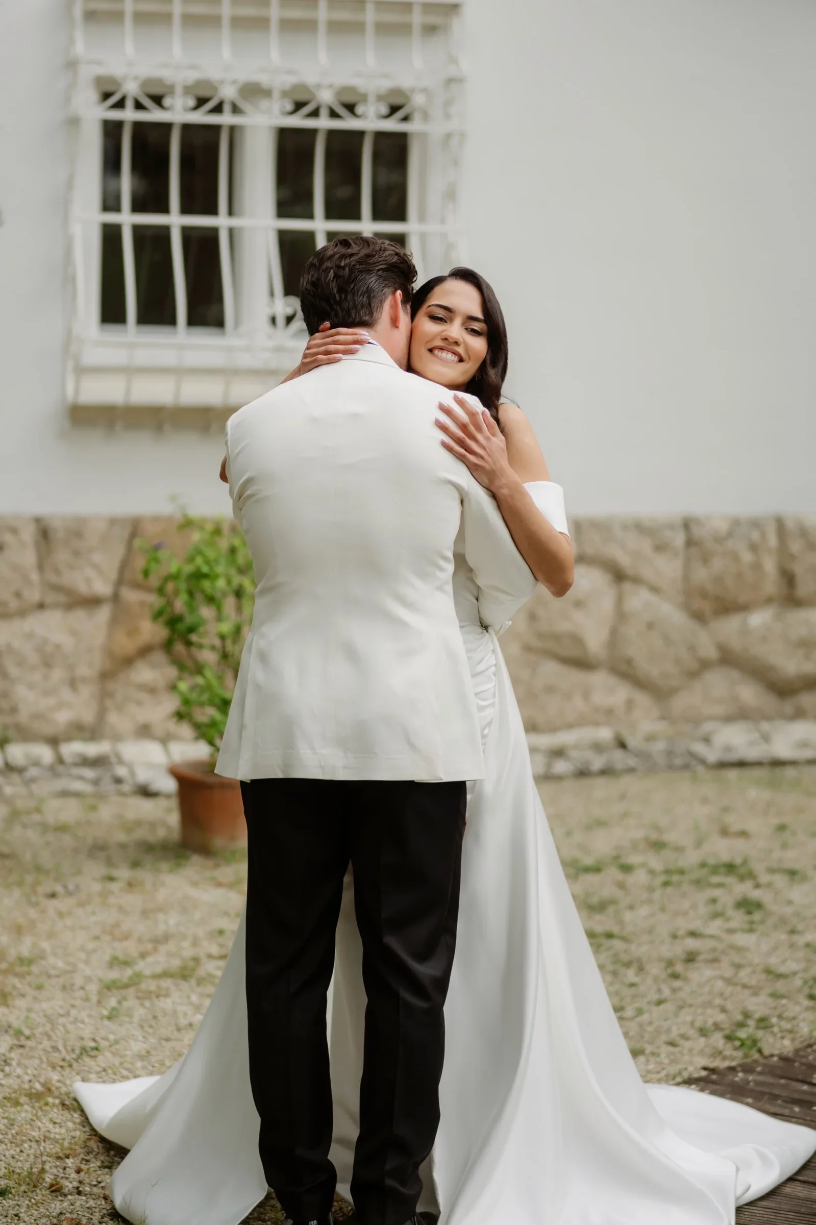 Groom in a white jacket hugging the bride in front of a stone wall