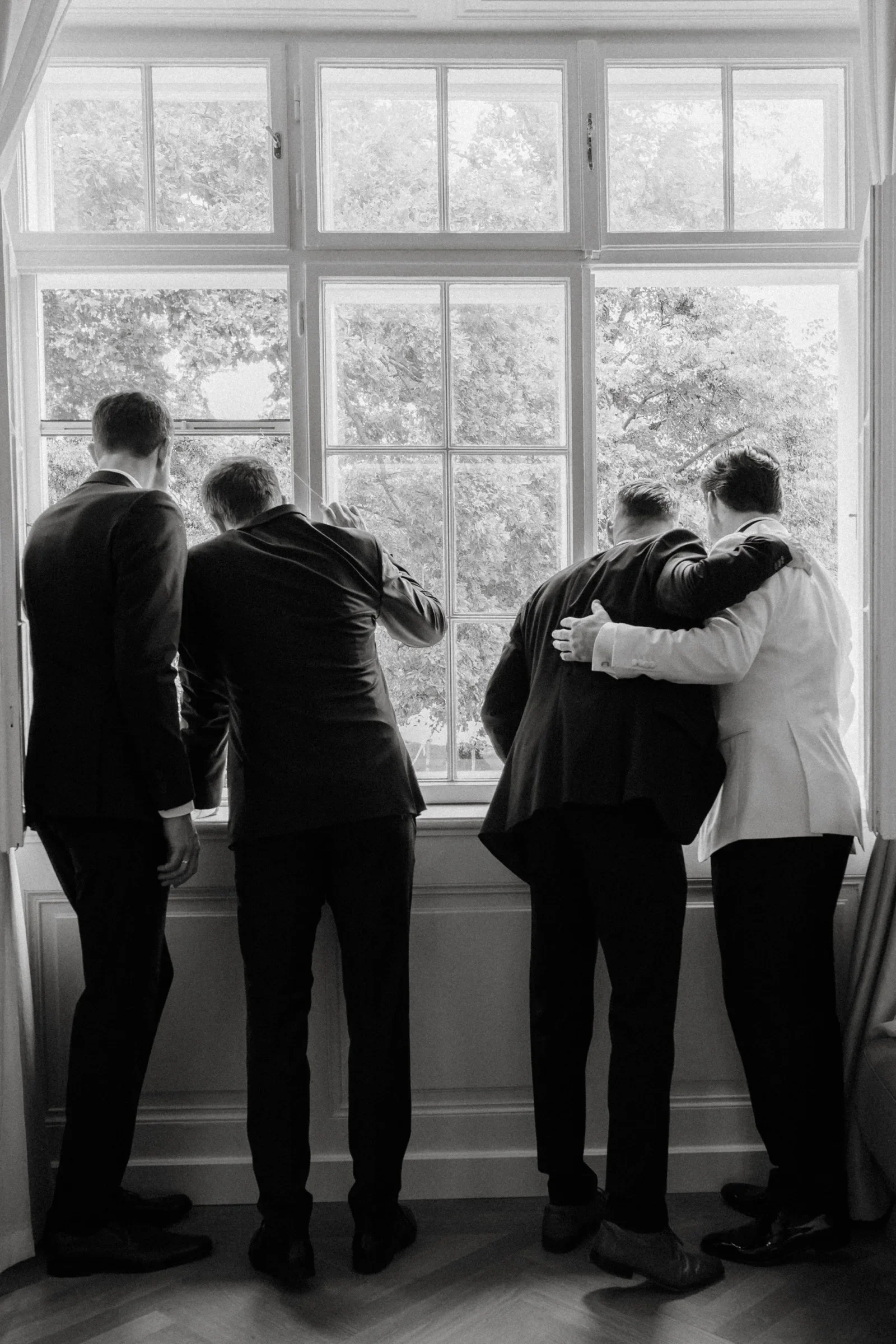 Black and white photo of the groom and groomsmen embracing in front of a tall window