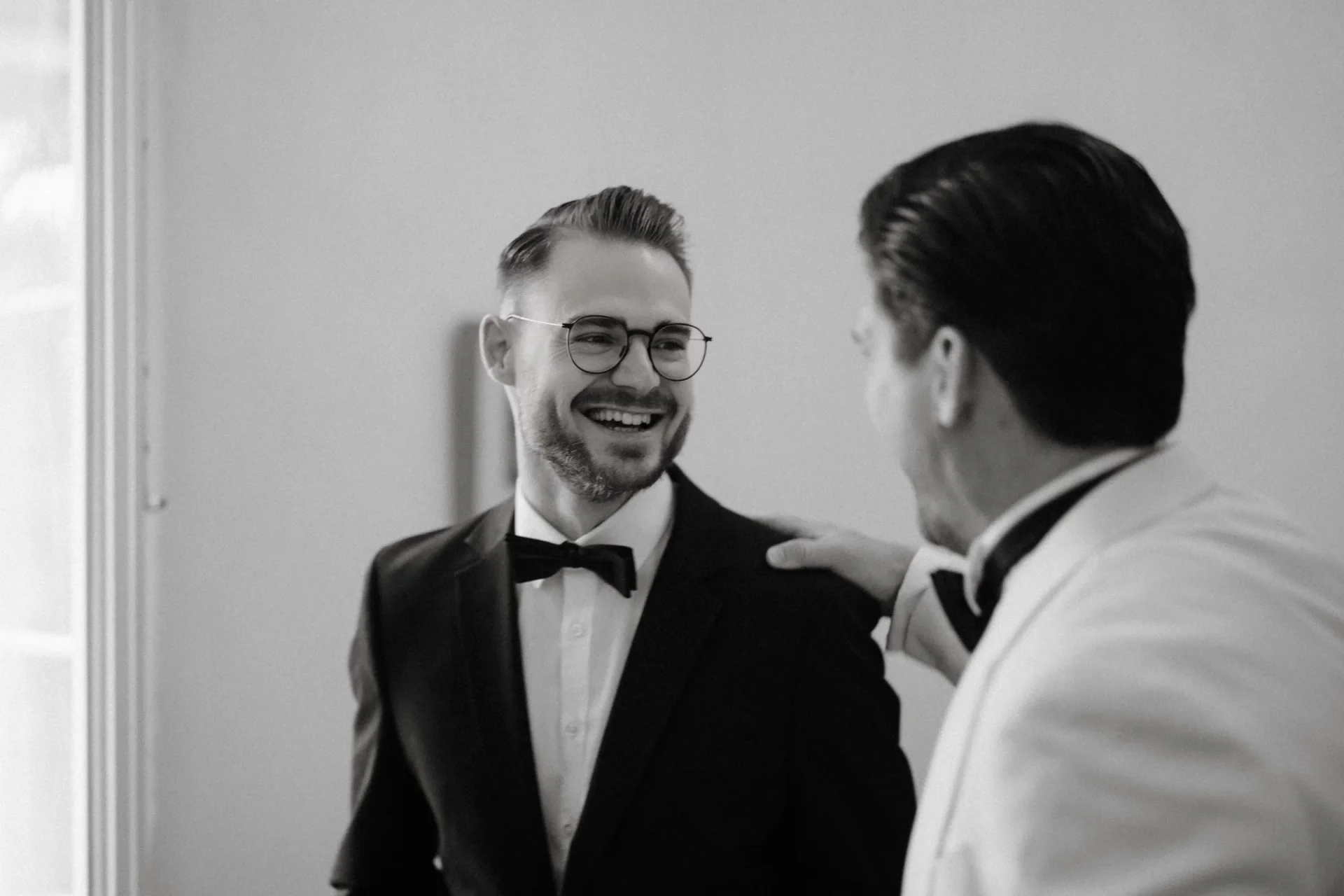 Black and white portrait of a smiling groom in glasses and a tuxedo while a friend pats his shoulder