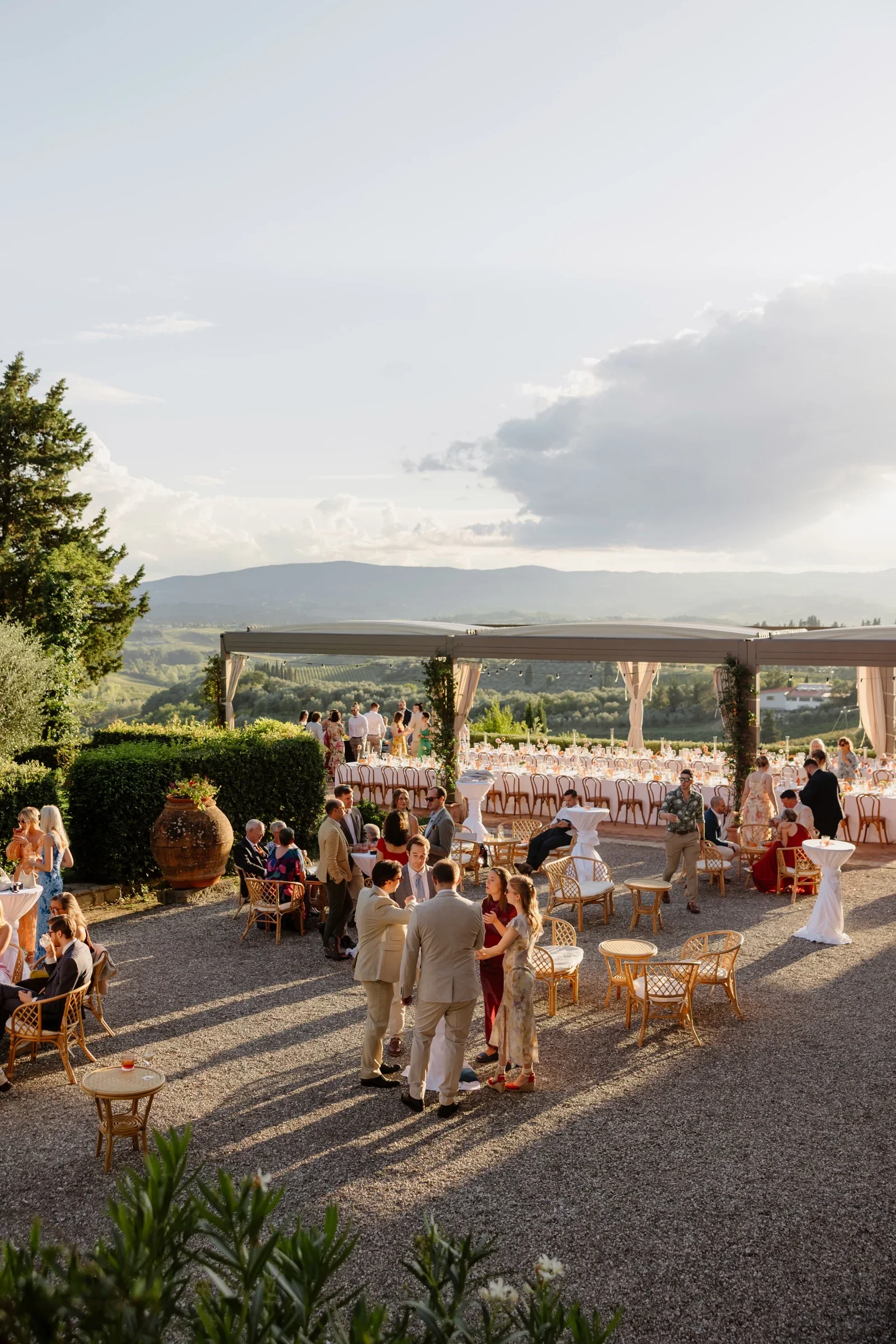 Aperitivo under the pergola at golden hour, guests scattered across the gravel terrace