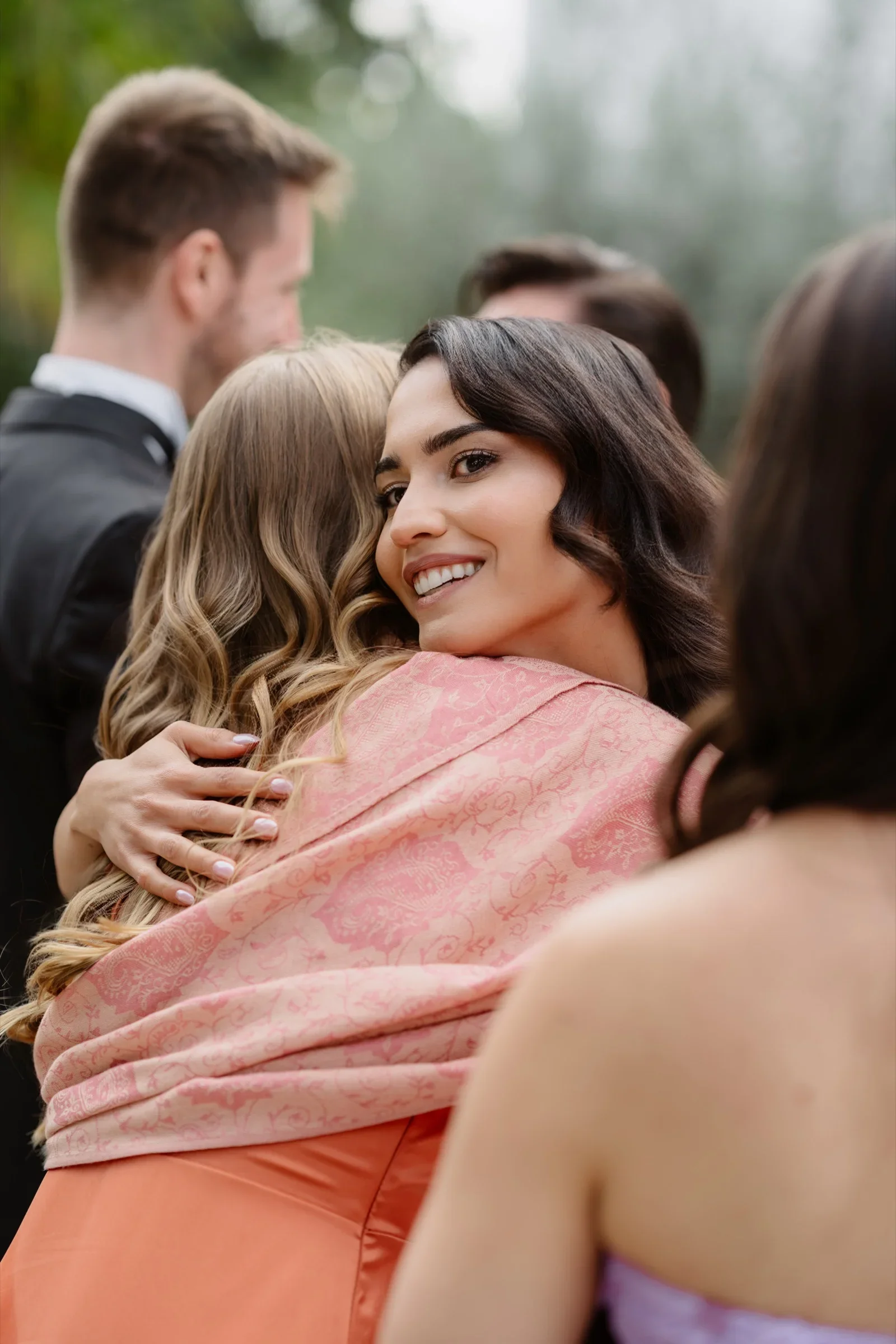 Bride in a veil smiling as she is hugged by a guest in a pink wrap
