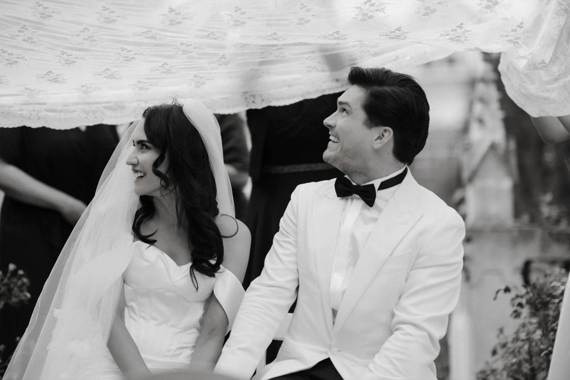 Bride and groom under a canopy, the groom in a white jacket and bow tie laughing