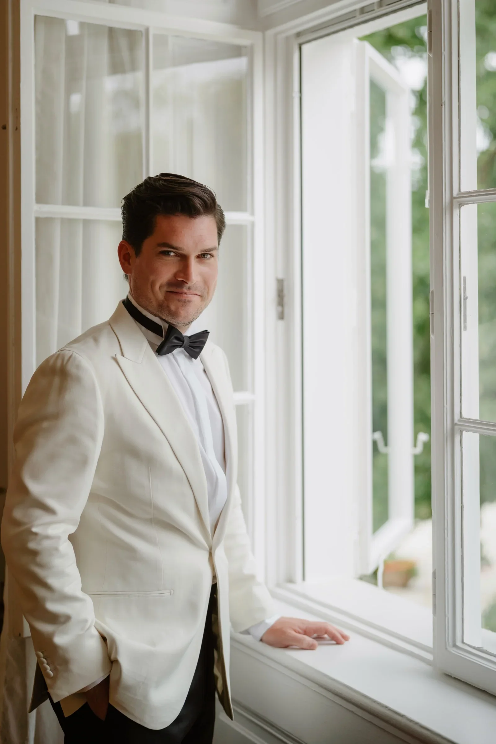 Groom in a white dinner jacket and bow tie standing by an open window