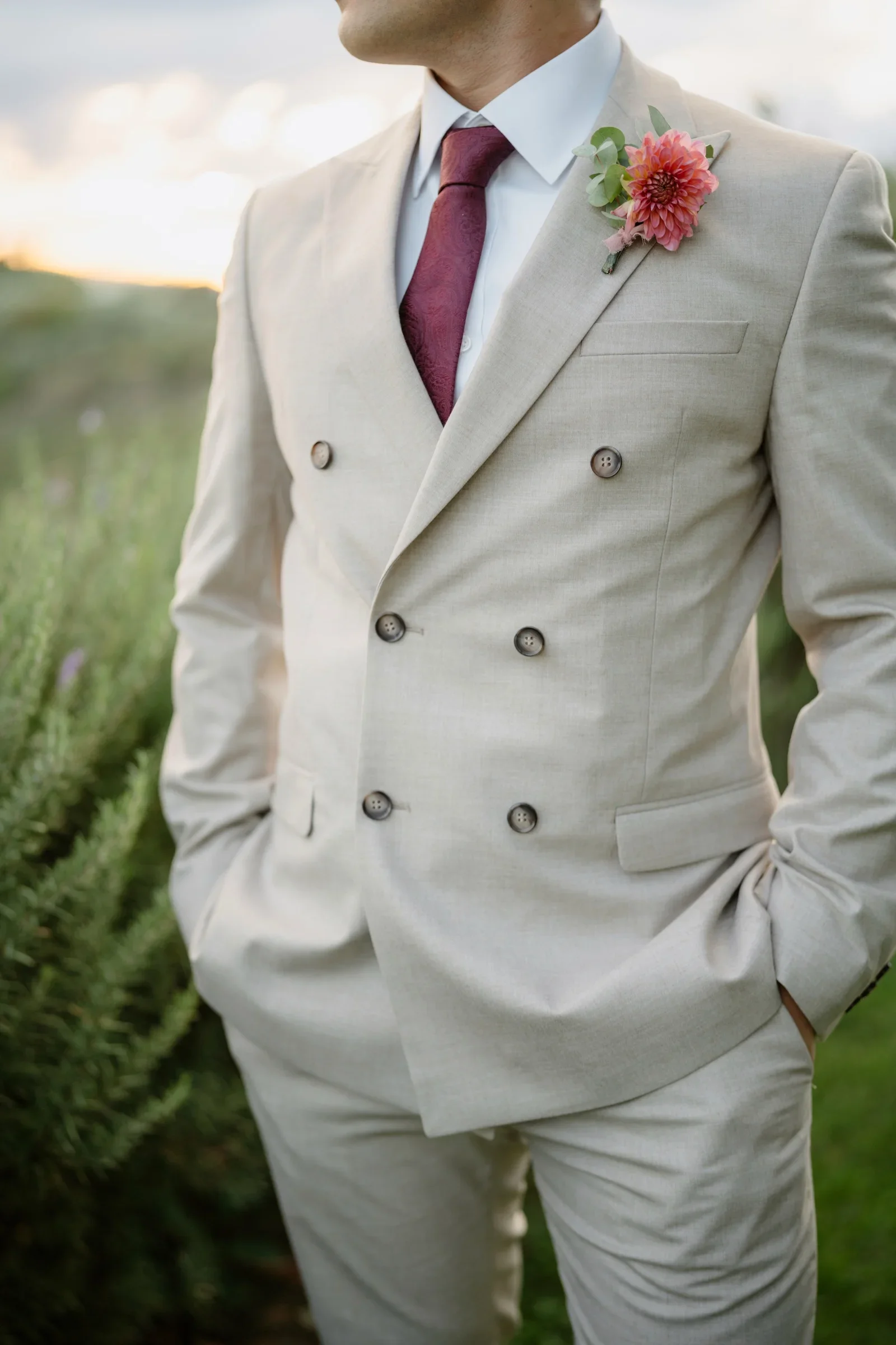 Close view of the groom's cream linen suit, burgundy tie and coral boutonniere
