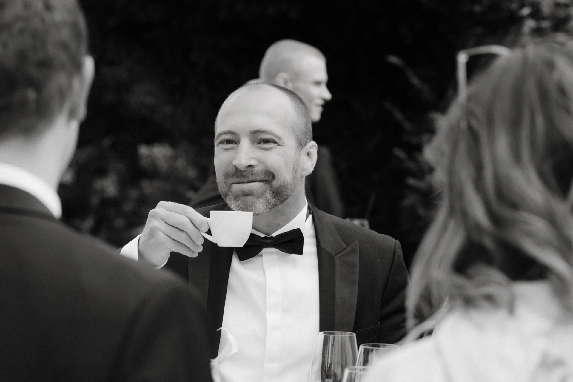 Black and white photo of a man in a bow tie smiling over a coffee cup
