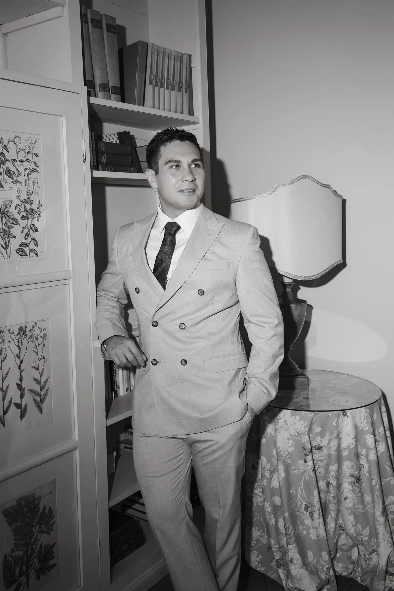 Black and white portrait of the groom in his cream suit, leaning against a bookcase