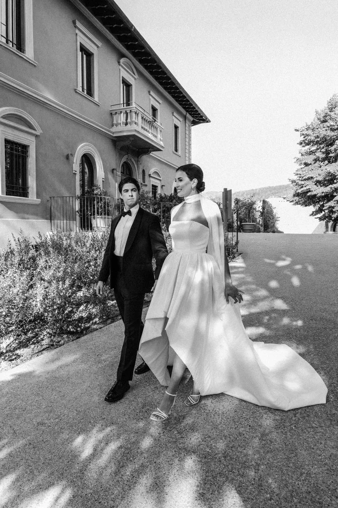 Bride and groom walking past an Italian villa, her train trailing behind — black and white